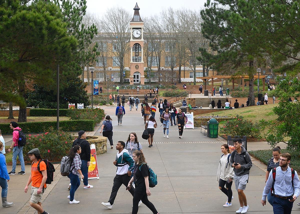 Many SHSU students traversing the middle of campus on a cloudy day. The clocktower stands in the background.